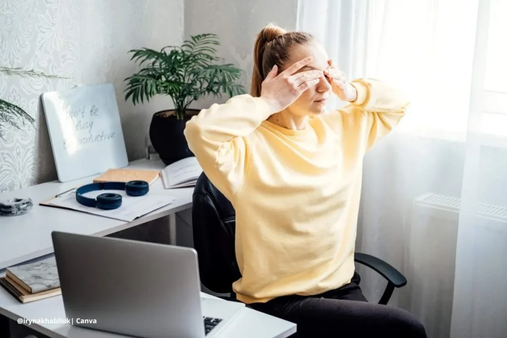 Mujer joven realizando ejercicios para los ojos frente a su escritorio con portátil abierto y luz natural entrando por la ventana.