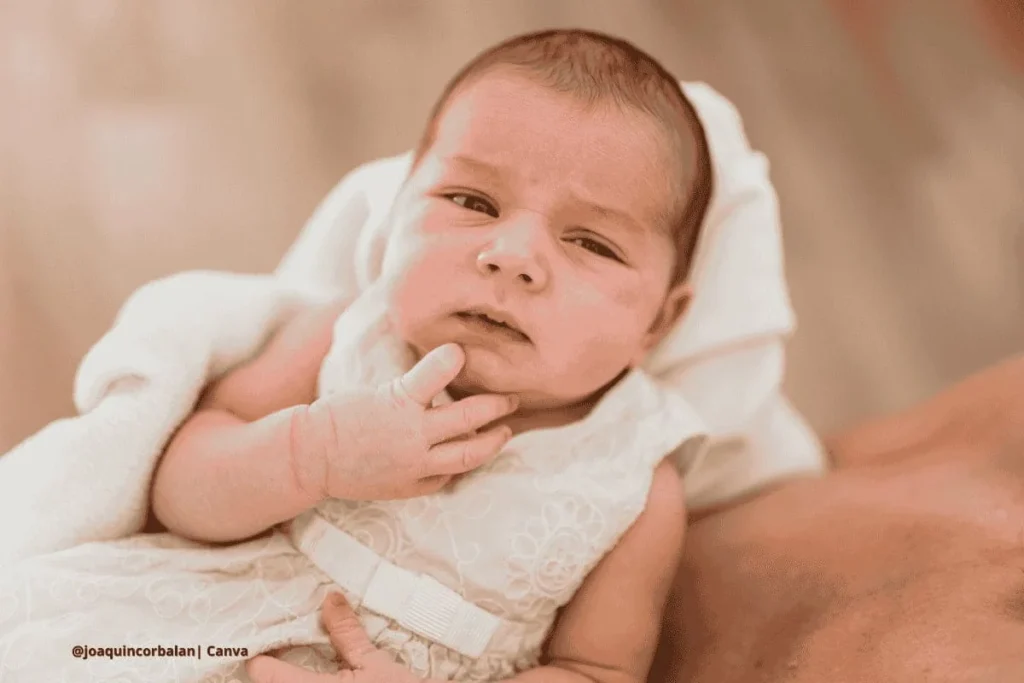 Bebé con expresión seria, vestido de blanco y recostado.