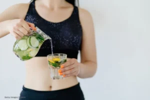 Mujer en ropa deportiva sirve agua saborizada con pepino y limón; importancia de la hidratación.